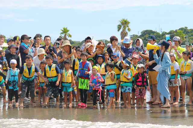 浜辺でカルタ取りのスタートラインに立つ子供たち。保護者はカルタを読み上げる酒井さんをスマホで撮影しています