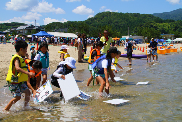浜辺近くの海辺に浮かべた大きなかるたのカードを取りに走る子供たち