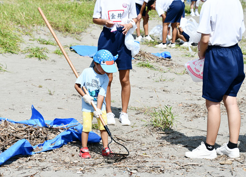 中学生のお姉さんの真似をして、熊手を使って流木を集めている男の子