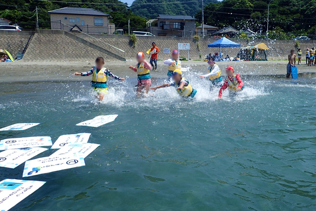 浜辺近くの海面に浮かぶ、大きなカルタのカードに向かって、海の中を水しぶきを上げつつ走る子供たち