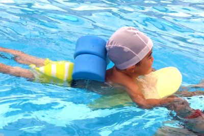 背中に子供用の浮き和代わりのスポンジ「ヘルパー」をつけて、ビート板を使って泳ぐ幼児